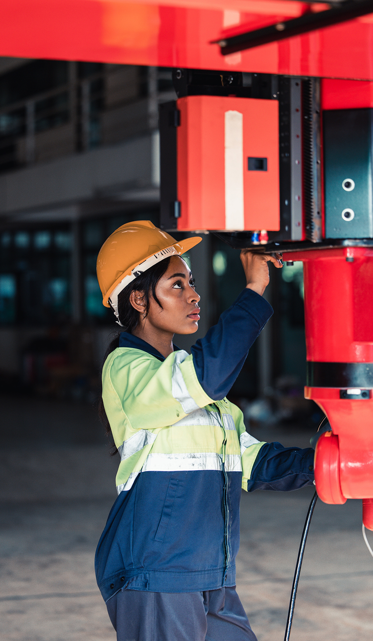 A female industrial worker in a yellow hard hat and safety jacket adjusts equipment on a large red machine inside a warehouse