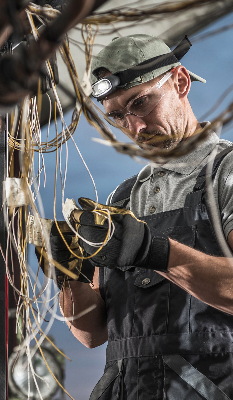 A technician in work overalls, safety glasses, and a headlamp focuses on wiring and complex electrical cables