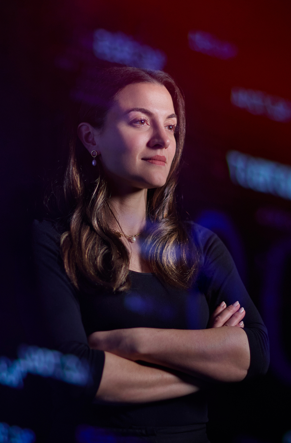 Confident Ann Kayis-Kumar with arms crossed, standing before a large, blurred financial screen.