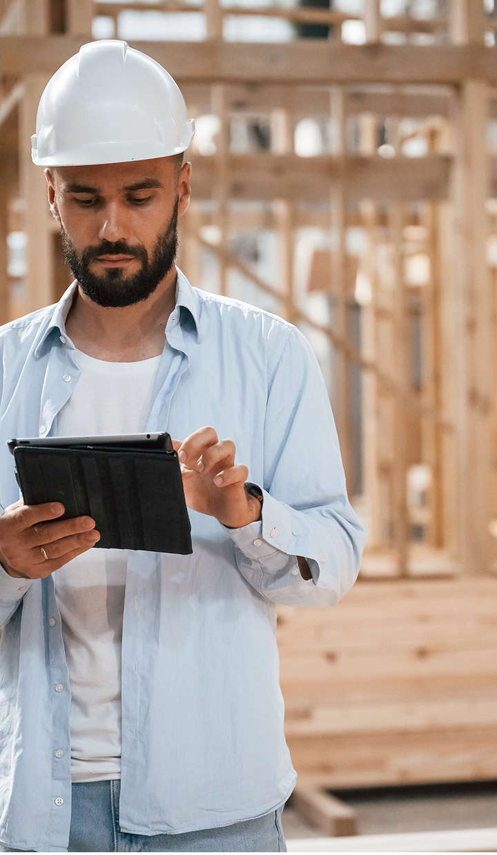 Construction foreman or wearing a white hard hat and blue shirt, reviewing plans on a digital tablet at a wooden construction site or building frame
