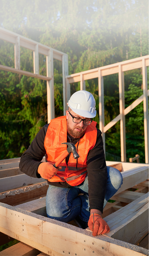 Construction worker in a hard hat and safety vest kneeling on a timber structure and hammering nails during a framing project