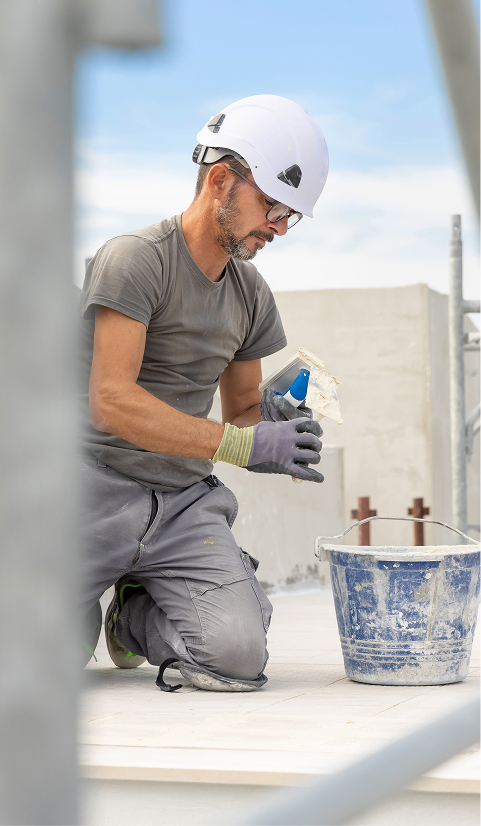Construction worker wearing a white hard hat kneeling on a job site and preparing materials beside a bucket