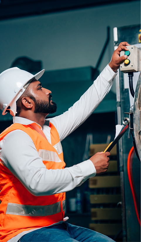 Factory technician in a hard hat and orange safety vest adjusting controls on industrial equipment