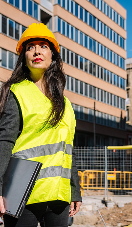 Female construction professional in a hard hat and high-visibility vest holding a laptop at an urban building site
