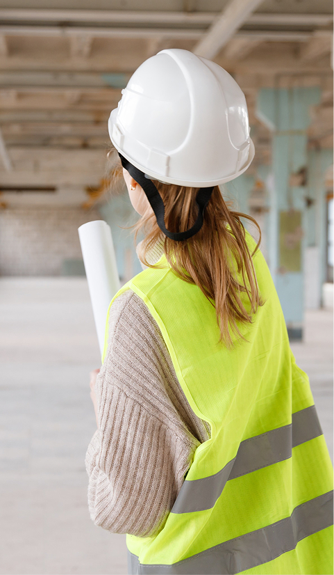 Female construction worker wearing a hard hat and high-visibility vest holding rolled building plans inside an unfinished structure