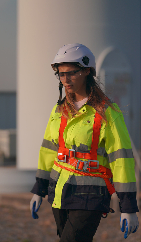 Female technician in high-visibility gear and safety harness walking near a wind turbine