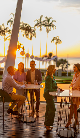 Group of people socialising at an outdoor venue during sunset, with palm trees and waterfront views in the background in Northern Territory