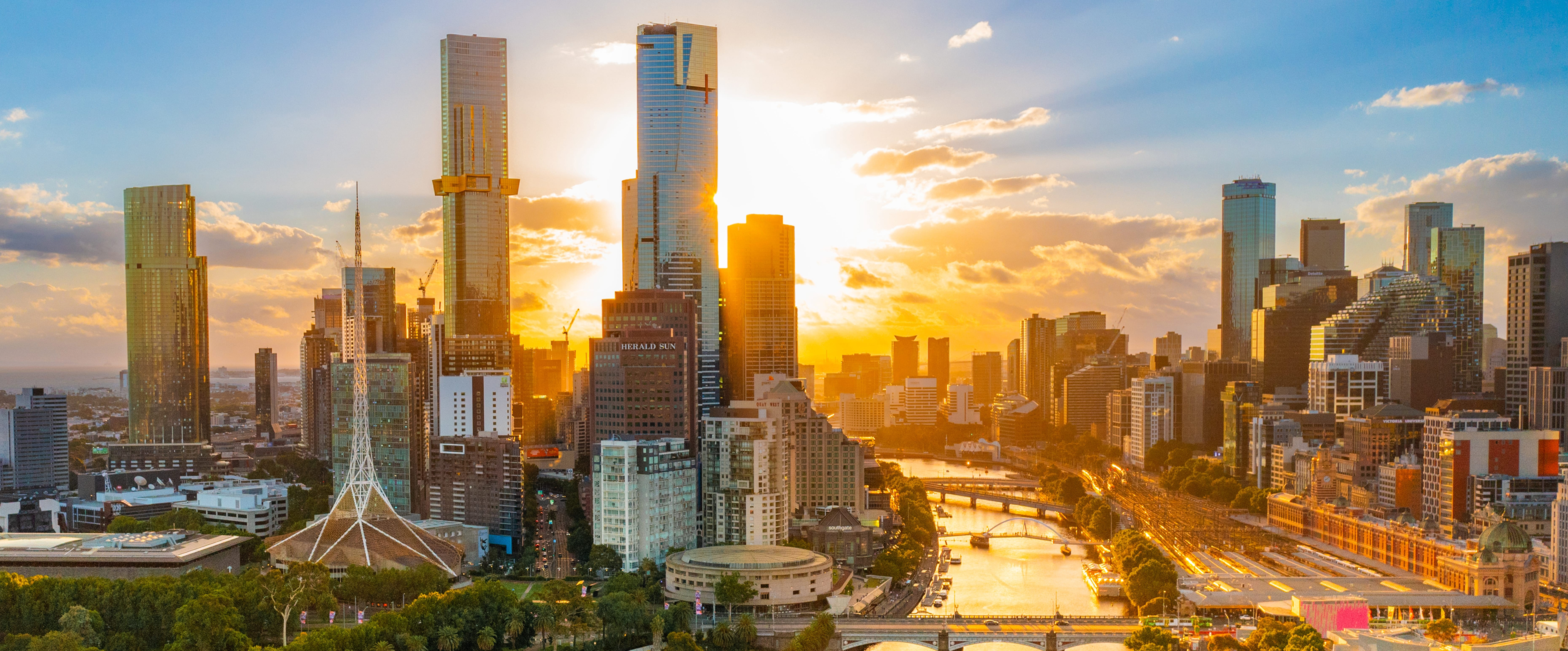 Sunset aerial of Melbourne CBD with Yarra River, tall buildings glowing in warm golden light.