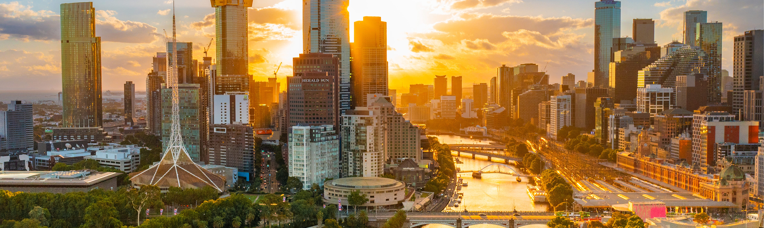 Sunset aerial of Melbourne CBD with Yarra River, tall buildings glowing in warm golden light.