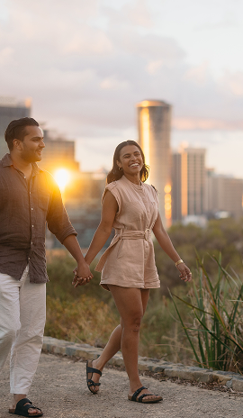 Smiling couple holding hands and walking outdoors at sunset with a city skyline in the background in Perth