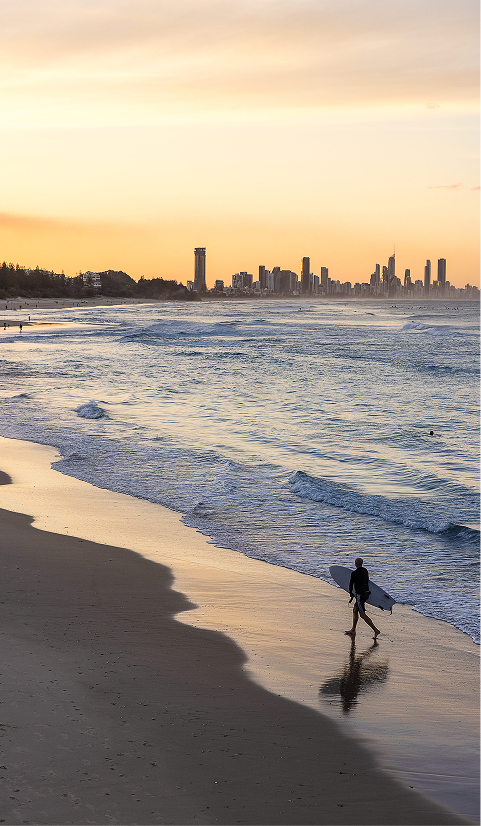 Surfer walking along a Gold Coast beach at sunset with the city skyline in the distance