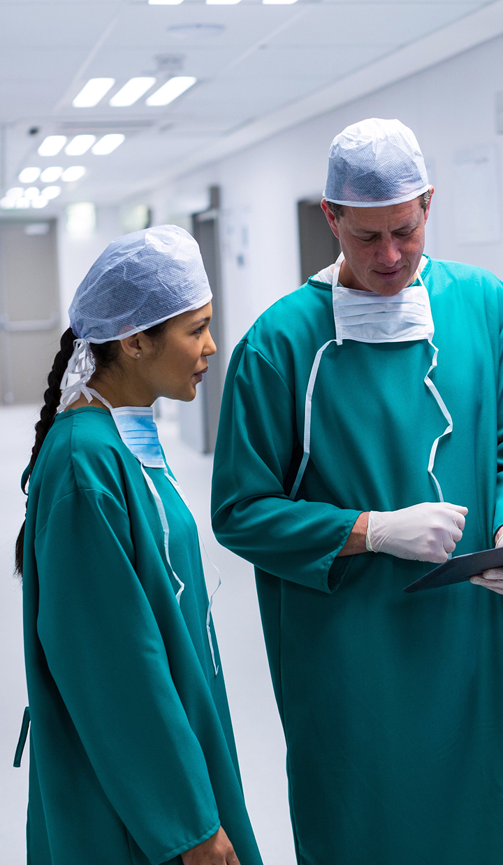 Two medical professionals wearing green surgical gowns, caps, and gloves are standing in a brightly lit hospital corridor, engaged in discussion.