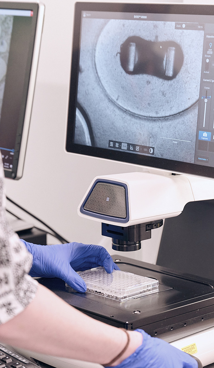 gloved hands operating microscope with monitor displaying magnified image