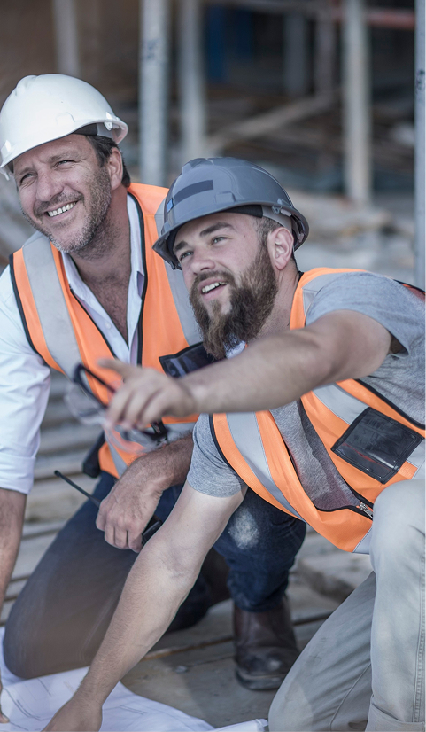 Two construction workers in hard hats and high-visibility vests smiling and reviewing building plans onsite