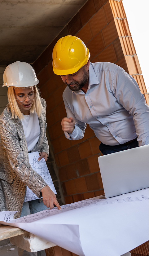 Two engineers in hard hats reviewing architectural plans with a laptop on a construction site