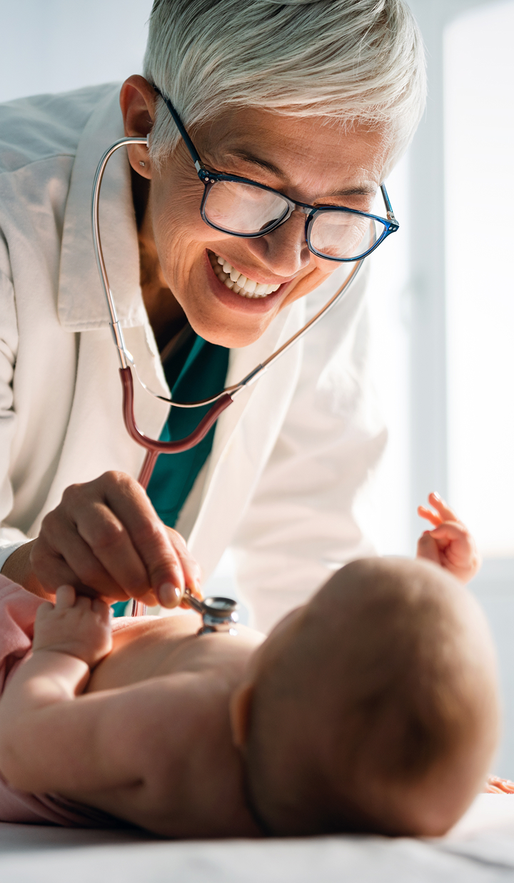 A healthcare professional wearing a white coat is using a stethoscope to examine an infant lying on an examination table.