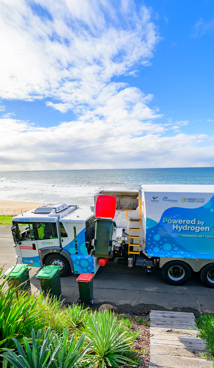 A hydrogen-powered truck collecting bins by the seaside under a blue sky.