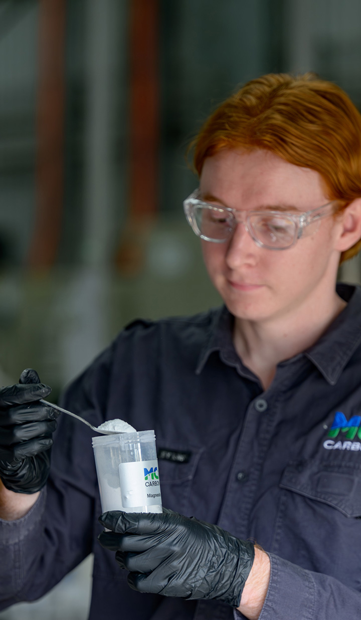 A person wearing safety glasses and gloves holding a container of laboratory powder.