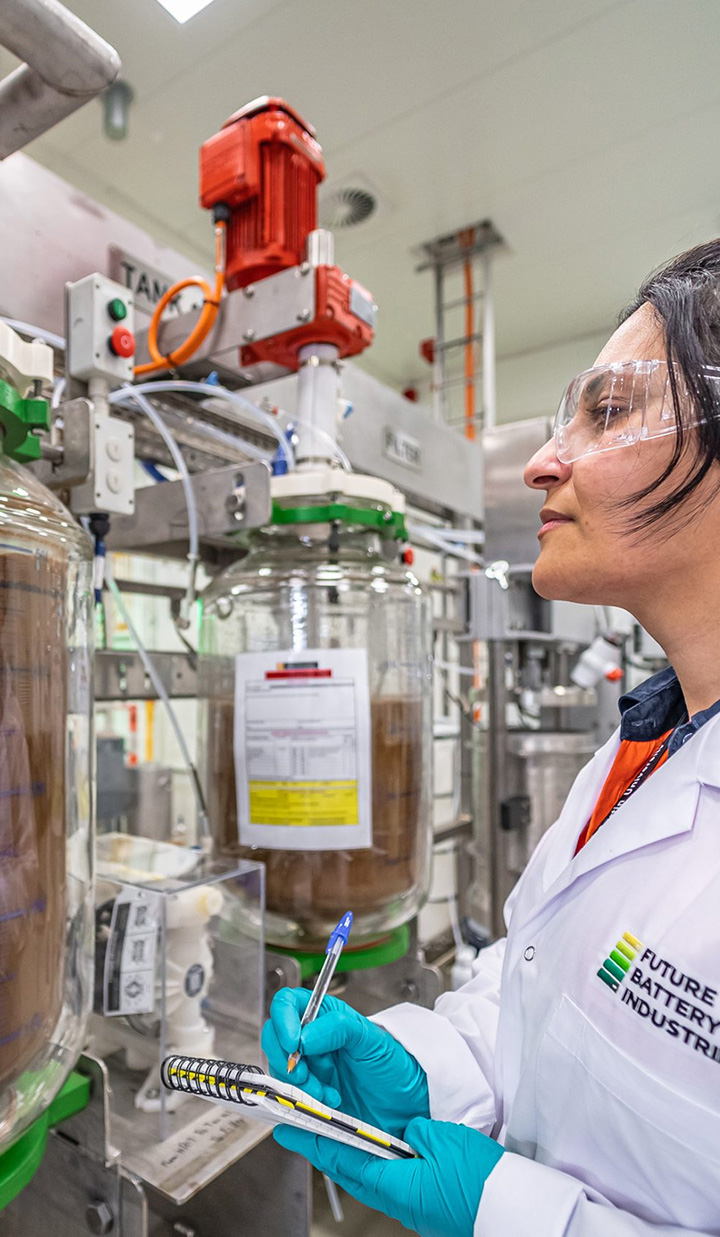 A woman from Future Battery Industries wearing PPE gear, inspecting a machine and taking notes.