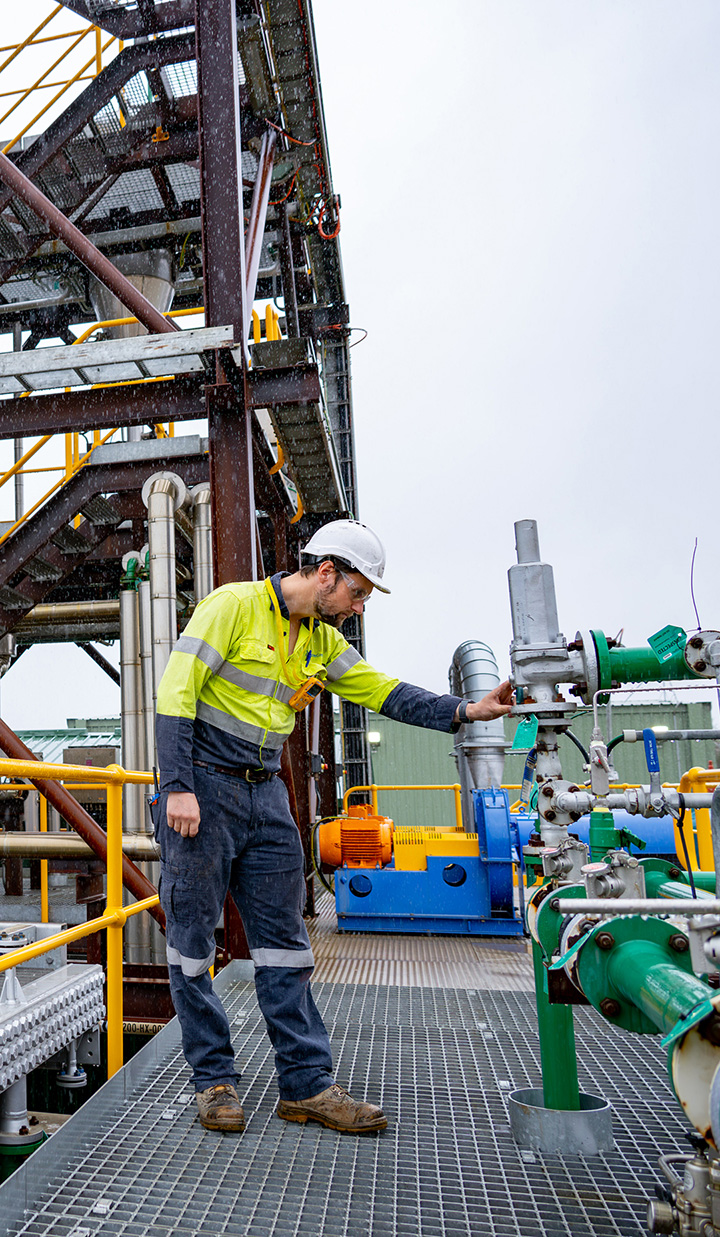 A worker in high-visibility clothing and a hard hat operating industrial machinery at an outdoor processing plant.