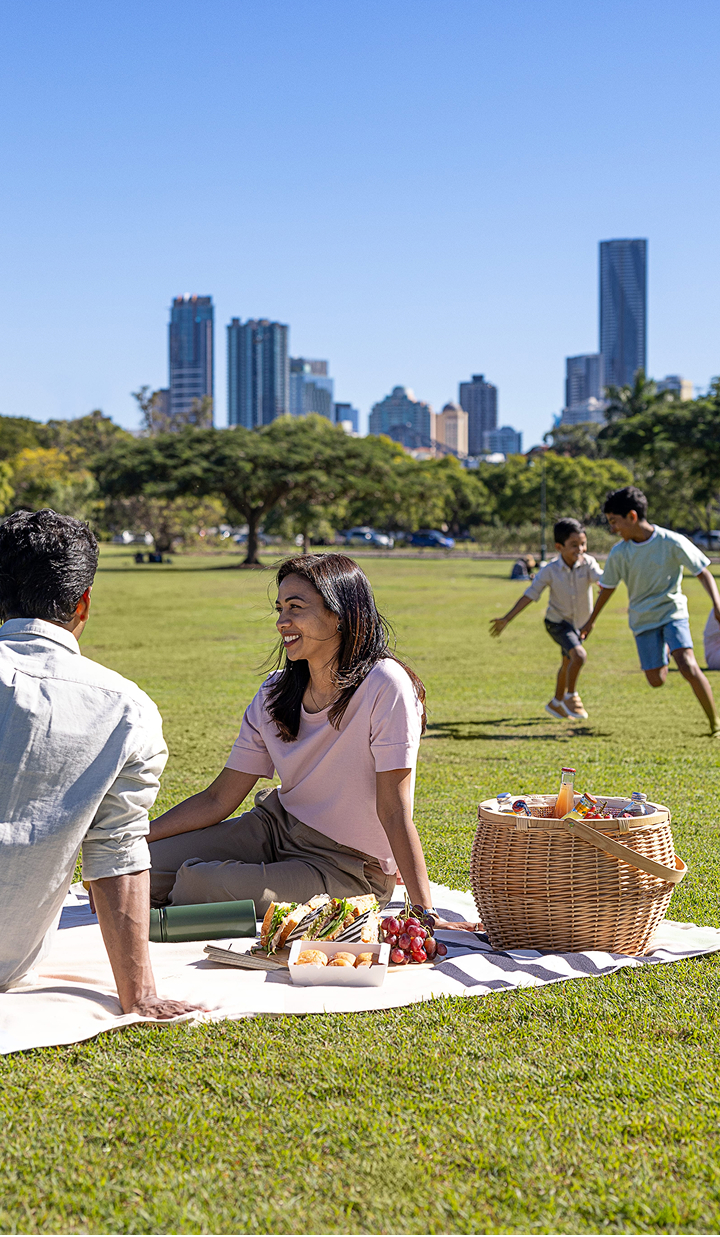 Family enjoys a picnic in a park as children play and the city skyline rises behind them.