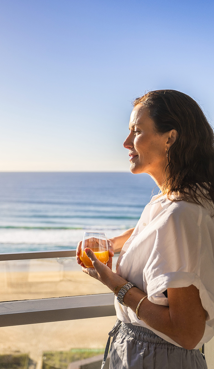 Woman with a glass of juice gazes at the beach in the early morning sun.