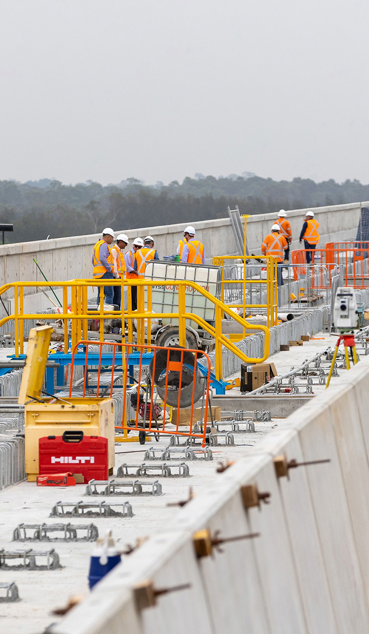 Workers in orange vests gathering for planning in a construction site, with barricades and scaffolds scattered along the path.