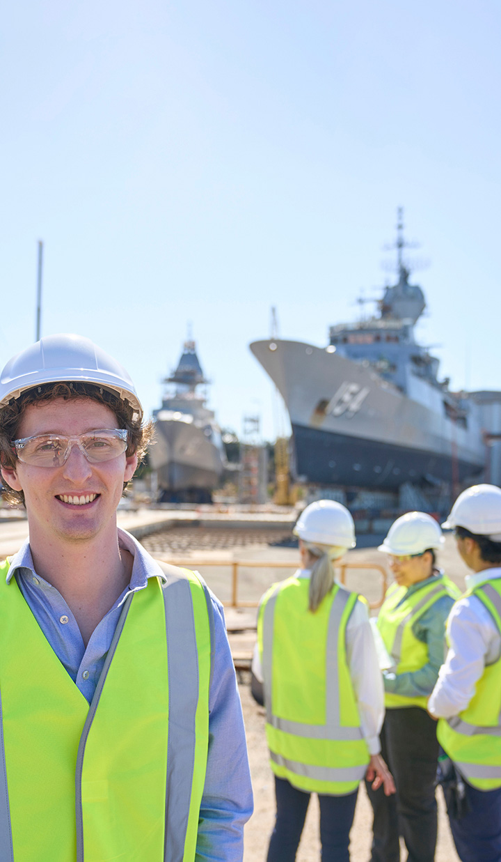 Smiling engineer in safety gear with ships in background.