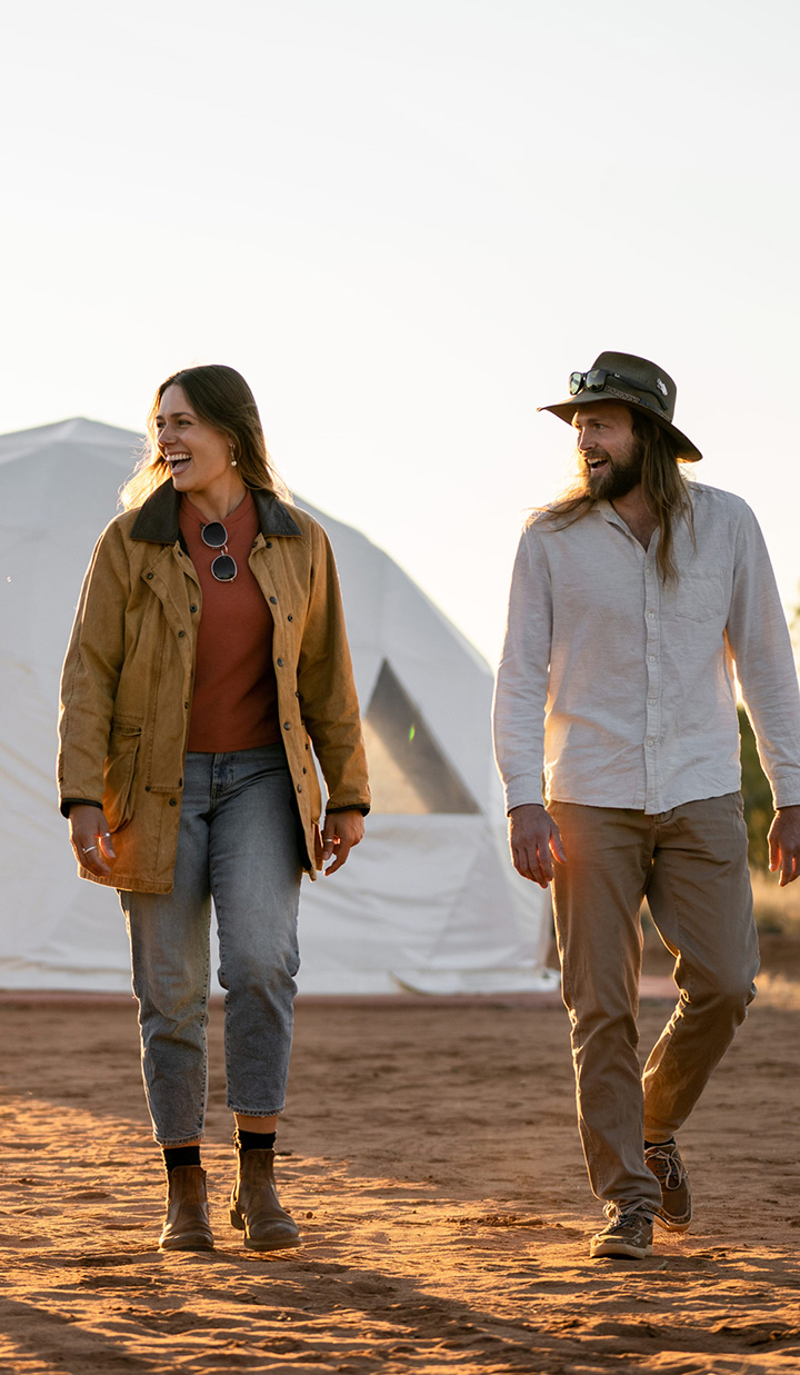Two people smiling walking on dirt with tents in background.