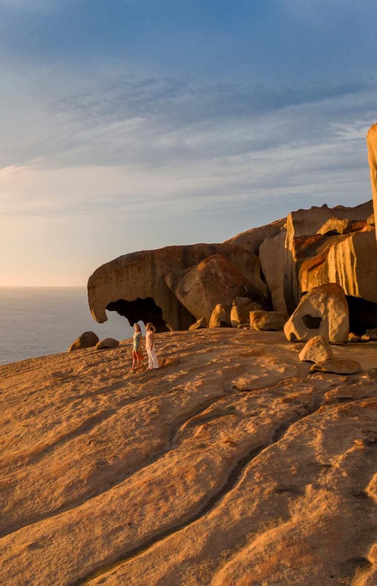 Two people standing on sunlit rock formations near dramatic coastal cliffs with the ocean in the background