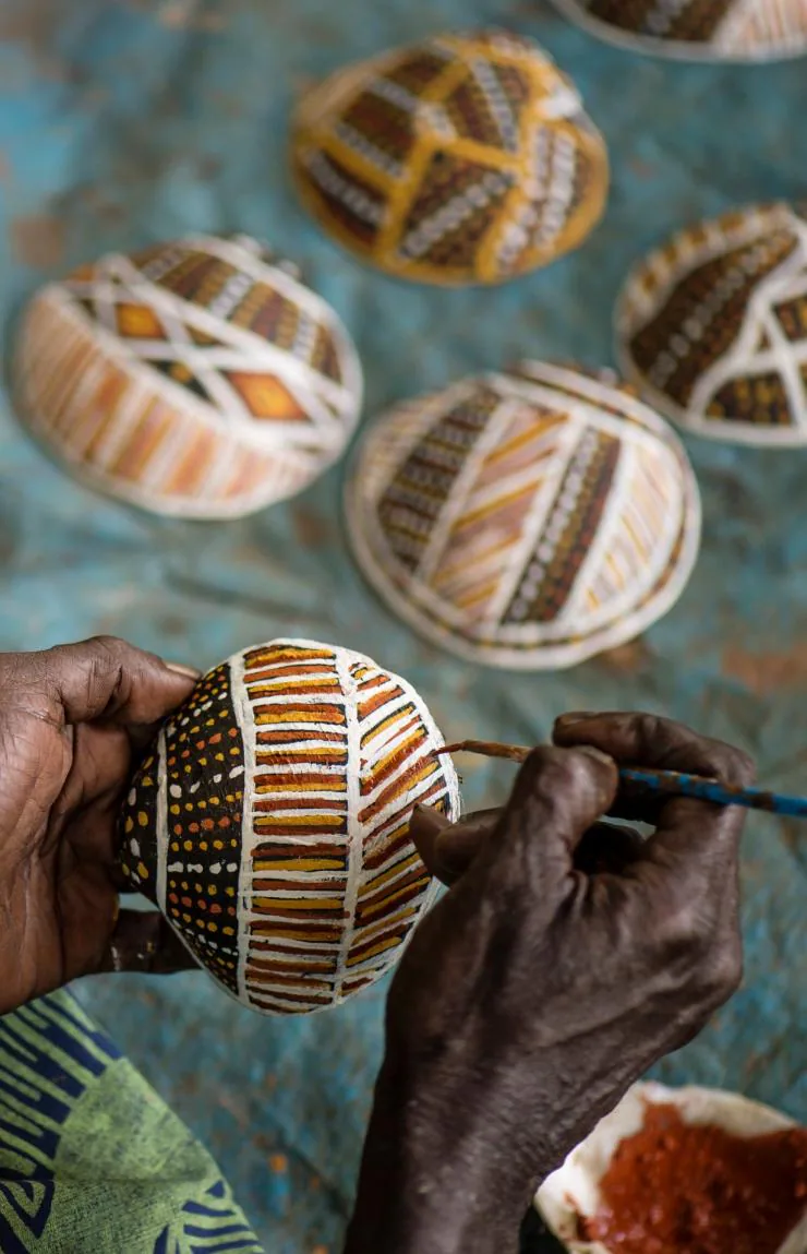 Hands painting traditional Indigenous Australian designs on a small, patterned object, with several finished pieces in the background