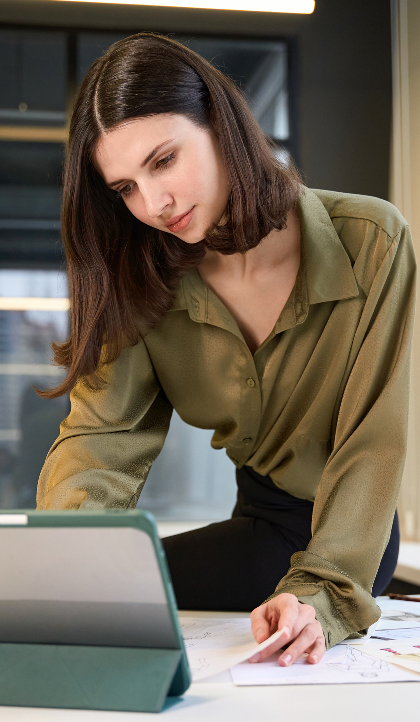 A person leaning over a table, working with a tablet and reviewing sketches or design papers in a modern office setting.