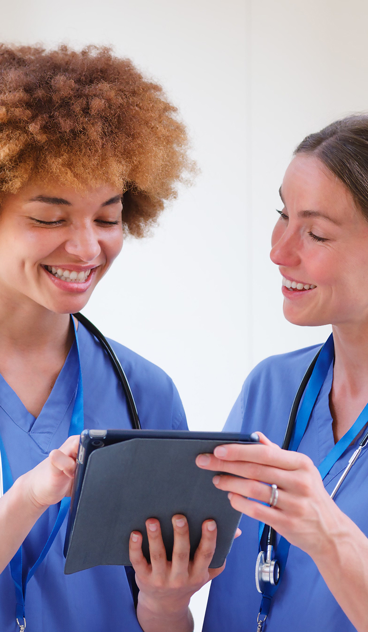 Two healthcare workers in scrubs review information together on a digital tablet.