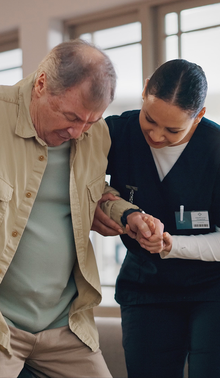 A caregiver supports an older adult as they stand and walk, holding their arm to provide stability in a bright indoor setting.