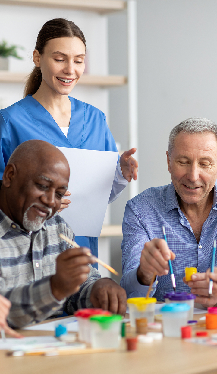 A group of aged adults sits at a table working on a painting activity, while a staff member in scrubs stands nearby holding a sheet of paper