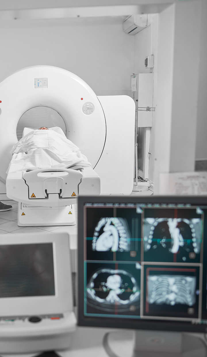A patient lies on a table being moved into a CT scanner, with medical imaging displayed on monitors in the foreground.