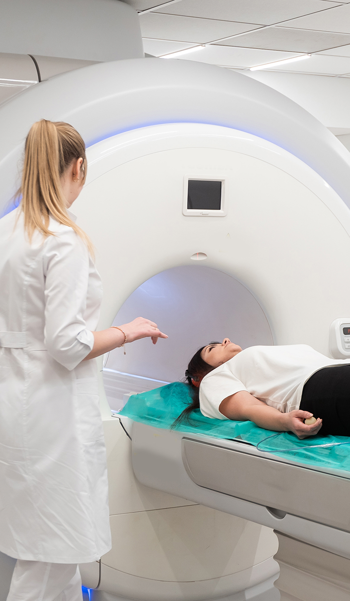 A healthcare professional stands beside a patient who is lying on a table and being positioned for an MRI scan in a medical imaging room.