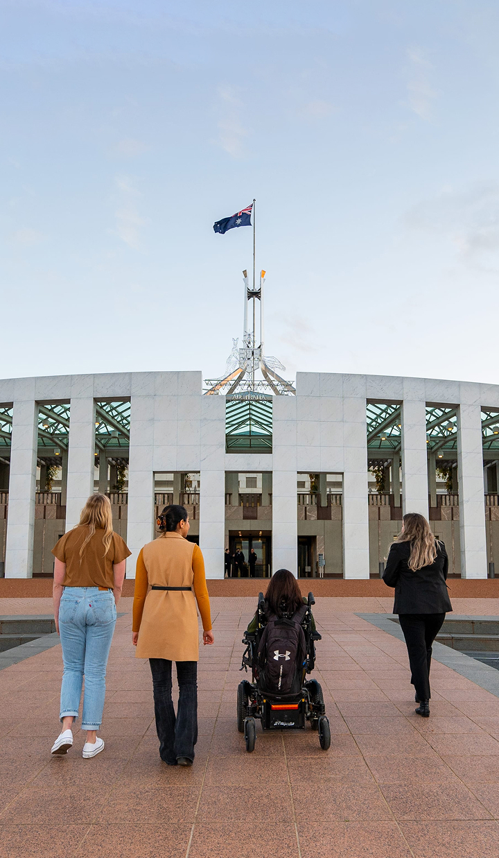 A group of four people, including one using a motorised wheelchair, walk toward the front entrance of Australia’s Parliament House in Canberra, with the Australian flag flying above the building.