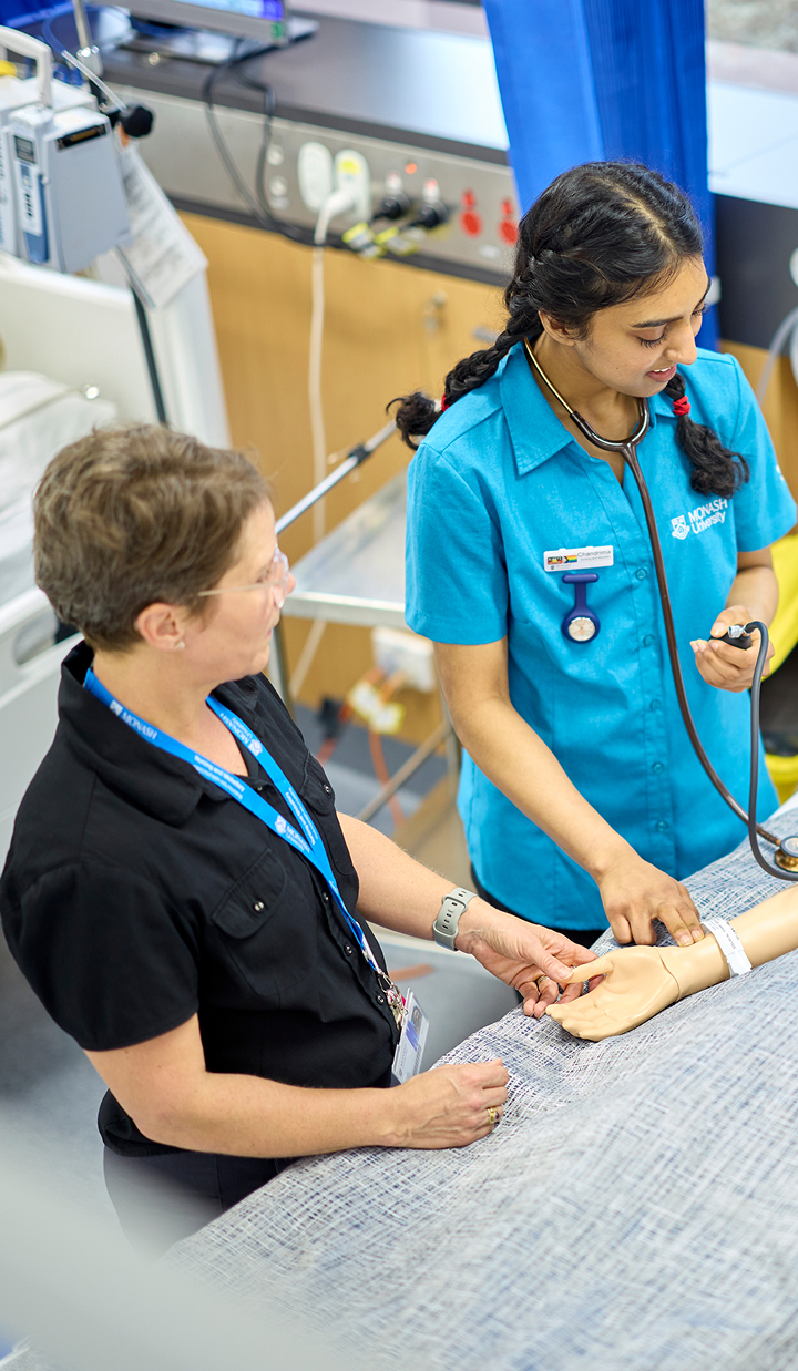 Two healthcare workers stand beside a patient’s bed in a clinical setting. One worker wearing a blue uniform uses a stethoscope to check the patient’s wrist, while the other worker observes and assists.