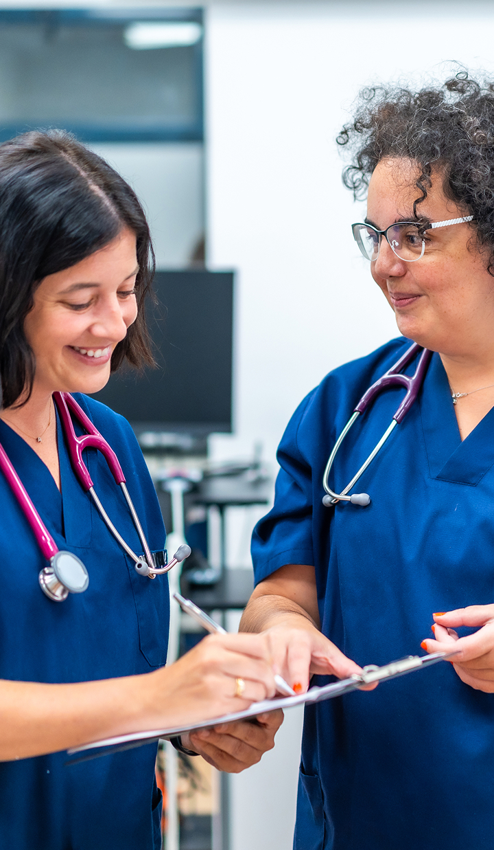 Two medical professionals wearing navy scrubs and stethoscopes review notes together on a clipboard in a clinical setting.