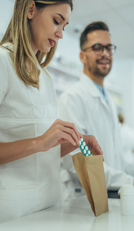 A pharmacy worker places a blister pack of pills into a small paper bag while another staff member stands in the background wearing a lab coat.