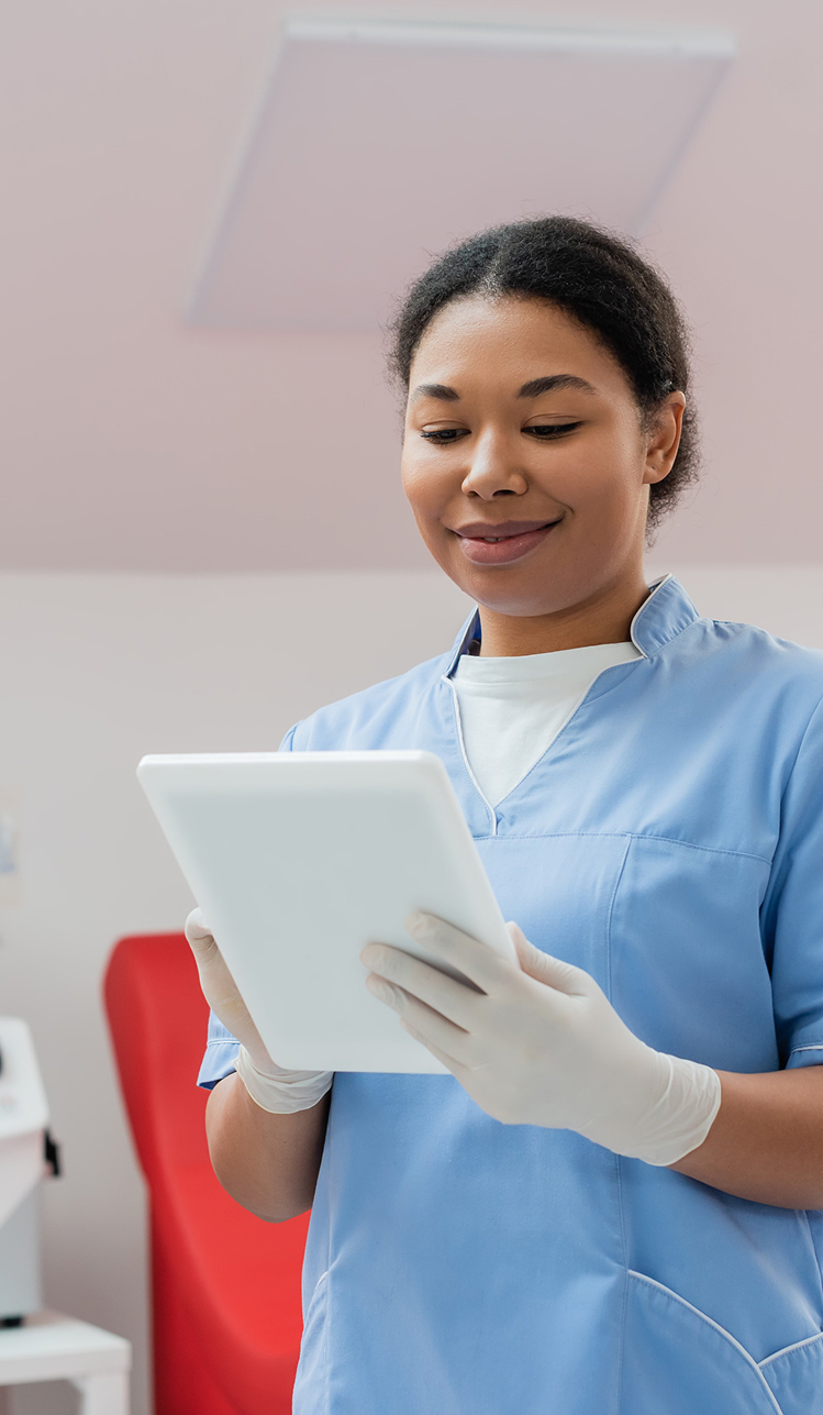 A healthcare worker wearing scrubs and gloves holds a digital tablet in a clinical setting