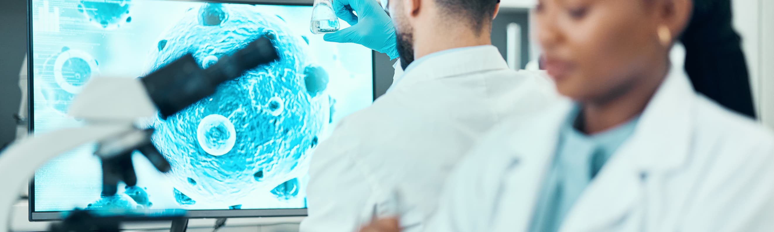 Scientists in a laboratory examine a flask and tablet beside a microscope image of cells displayed on a screen
