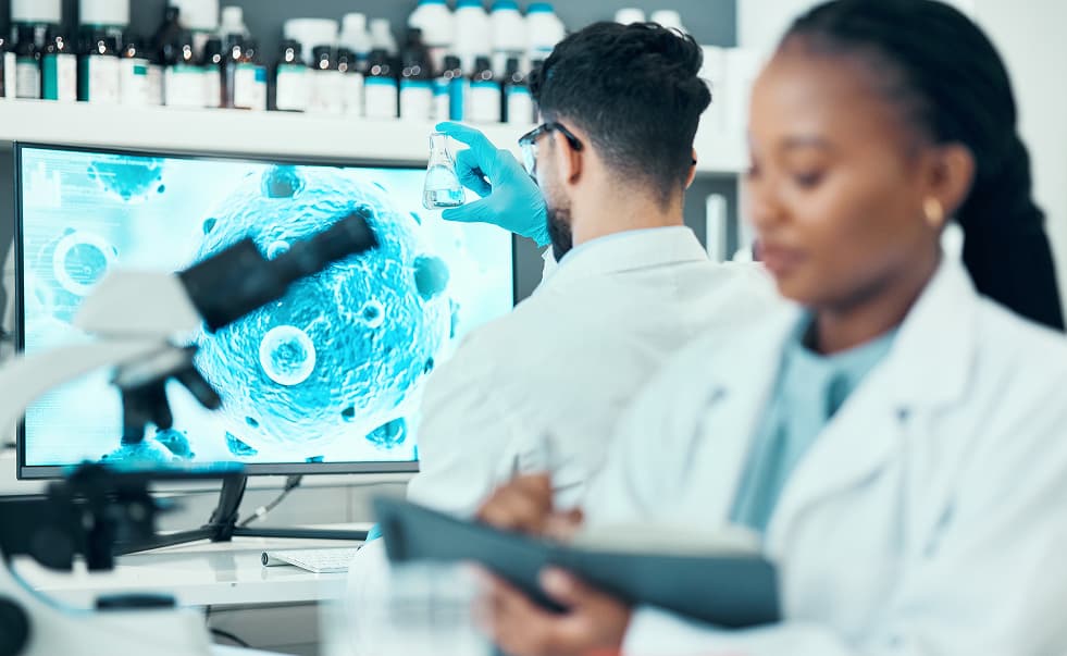 Scientists in a laboratory examine a flask and tablet beside a microscope image of cells displayed on a screen