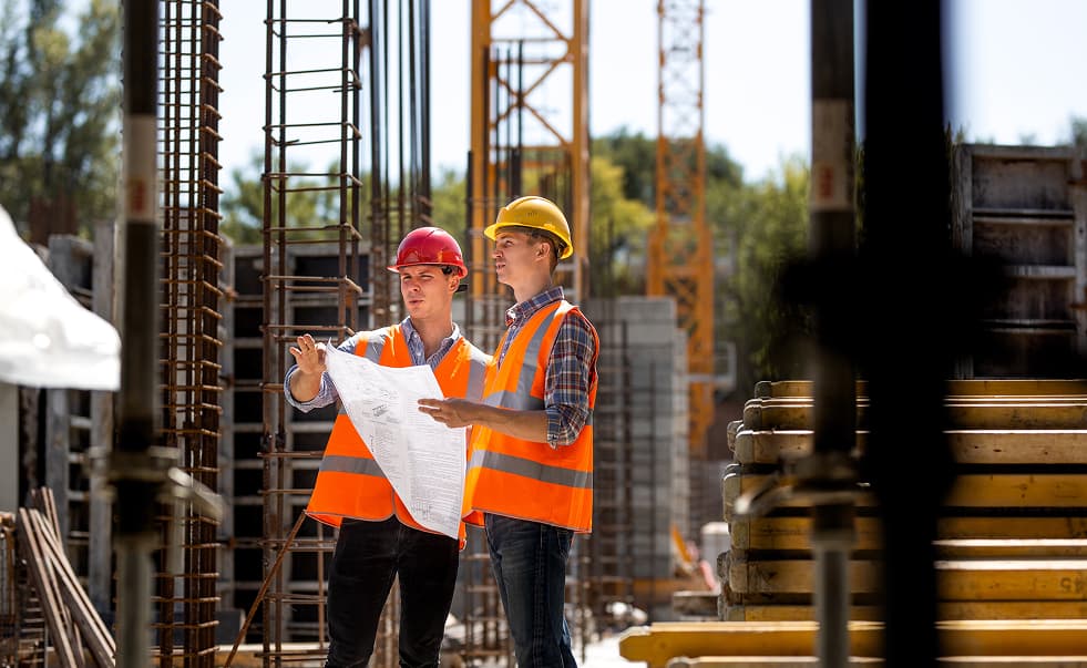 Two construction workers wearing safety vests and hard hats review blueprints at a building site with cranes and materials in the background.