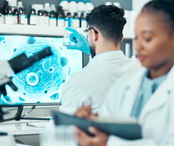 Scientists in a laboratory examine a flask and tablet beside a microscope image of cells displayed on a screen.
