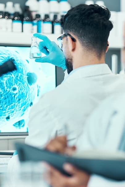 Scientists in a laboratory examine a flask and tablet beside a microscope image of cells displayed on a screen