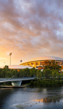 Adelaide Oval glowing at sunset with the riverfront and footbridge in the foreground