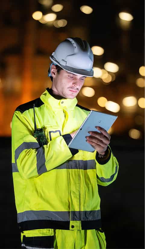 Construction worker in high-visibility gear and a hard hat using a tablet during night operations
