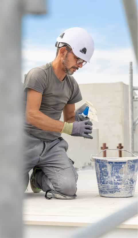 Construction worker wearing a white hard hat kneeling on a job site and preparing materials beside a bucket